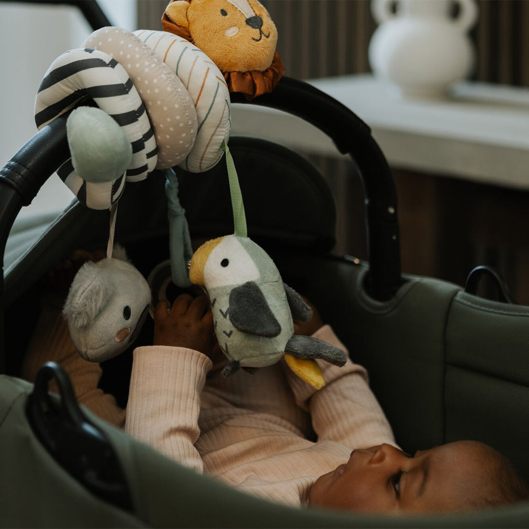 A baby in a pink outfit lies in a green stroller, reaching for the Animal Adventures Pram Spiral Activity Toy with soft hanging animal shapes—a lion, bird, and mouse—attached to the handle as a fun sensory stroller toy.