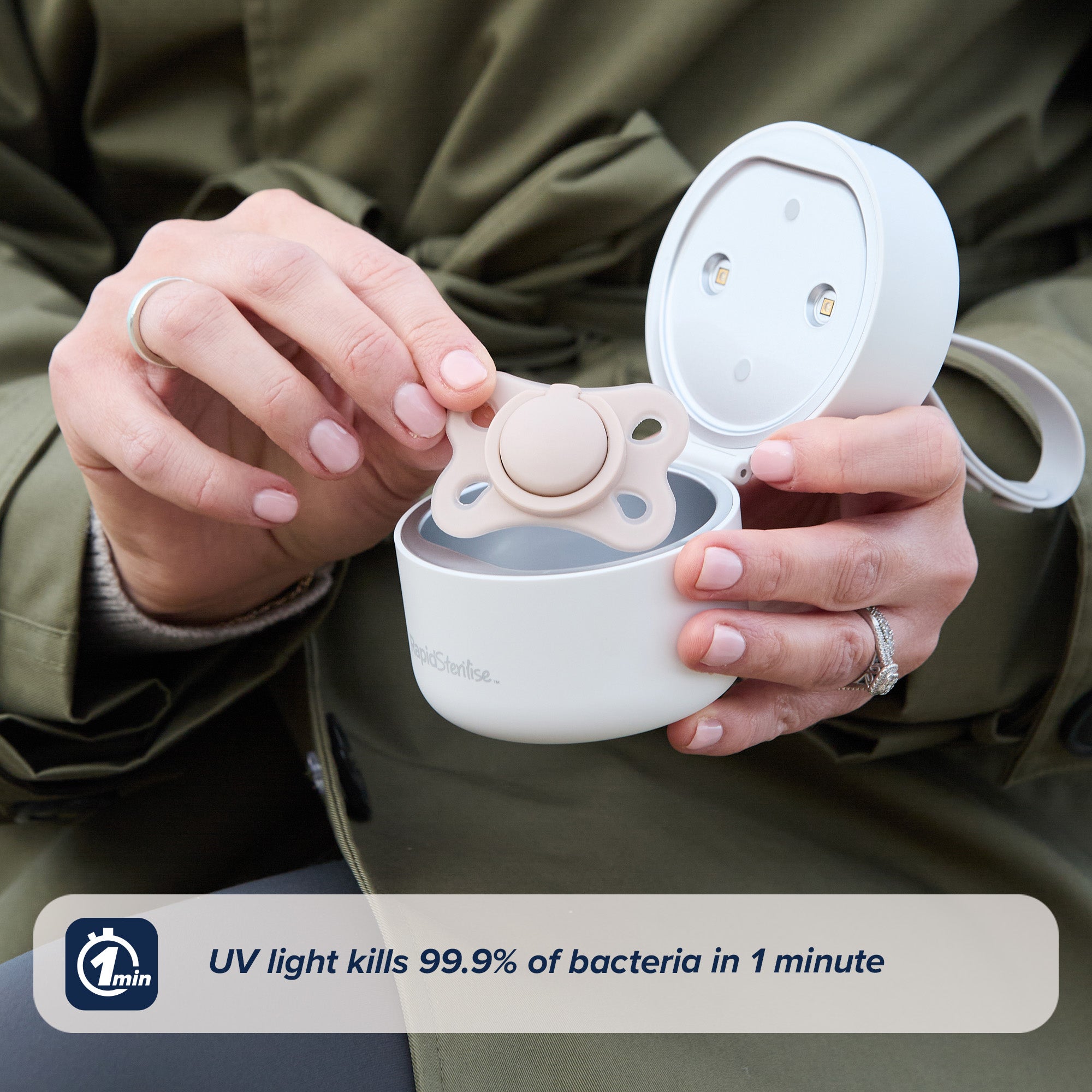 A person in a green coat puts a pacifier into the round, white RapidSterilise UV Soother Steriliser. Text reads "USB-powered UV kills 99.9% of bacteria in 1 minute," with a rapid sterilising claim and a 1-minute icon shown.