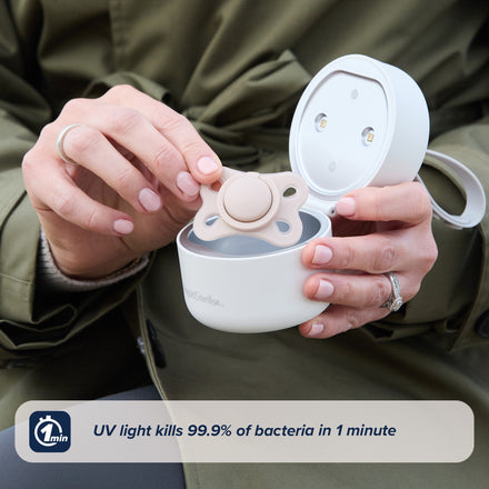 A person in a green coat puts a pacifier into the round, white RapidSterilise UV Soother Steriliser. Text reads "USB-powered UV kills 99.9% of bacteria in 1 minute," with a rapid sterilising claim and a 1-minute icon shown.