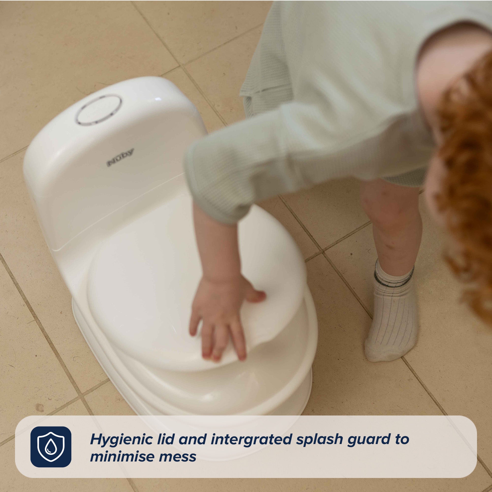 A young child in green clothes and white socks closes the lid of a white My Real Mini Toilet Potty on a tiled floor. Caption: Hygienic lid and integrated splash guard help minimise mess.