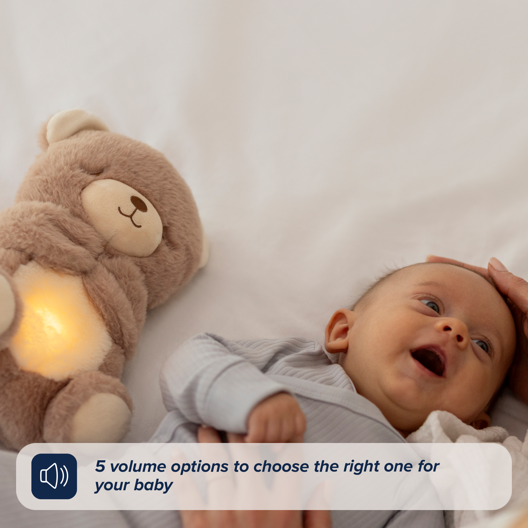 A smiling baby lies on a bed beside the glowing Biscuit Bear Calming Comforter as a hand gently touches the baby's head. Text: 5 volume options let you choose the perfect sleep aid setting for your little one.