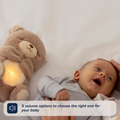 A smiling baby lies on a bed beside the glowing Biscuit Bear Calming Comforter as a hand gently touches the baby's head. Text: 5 volume options let you choose the perfect sleep aid setting for your little one.