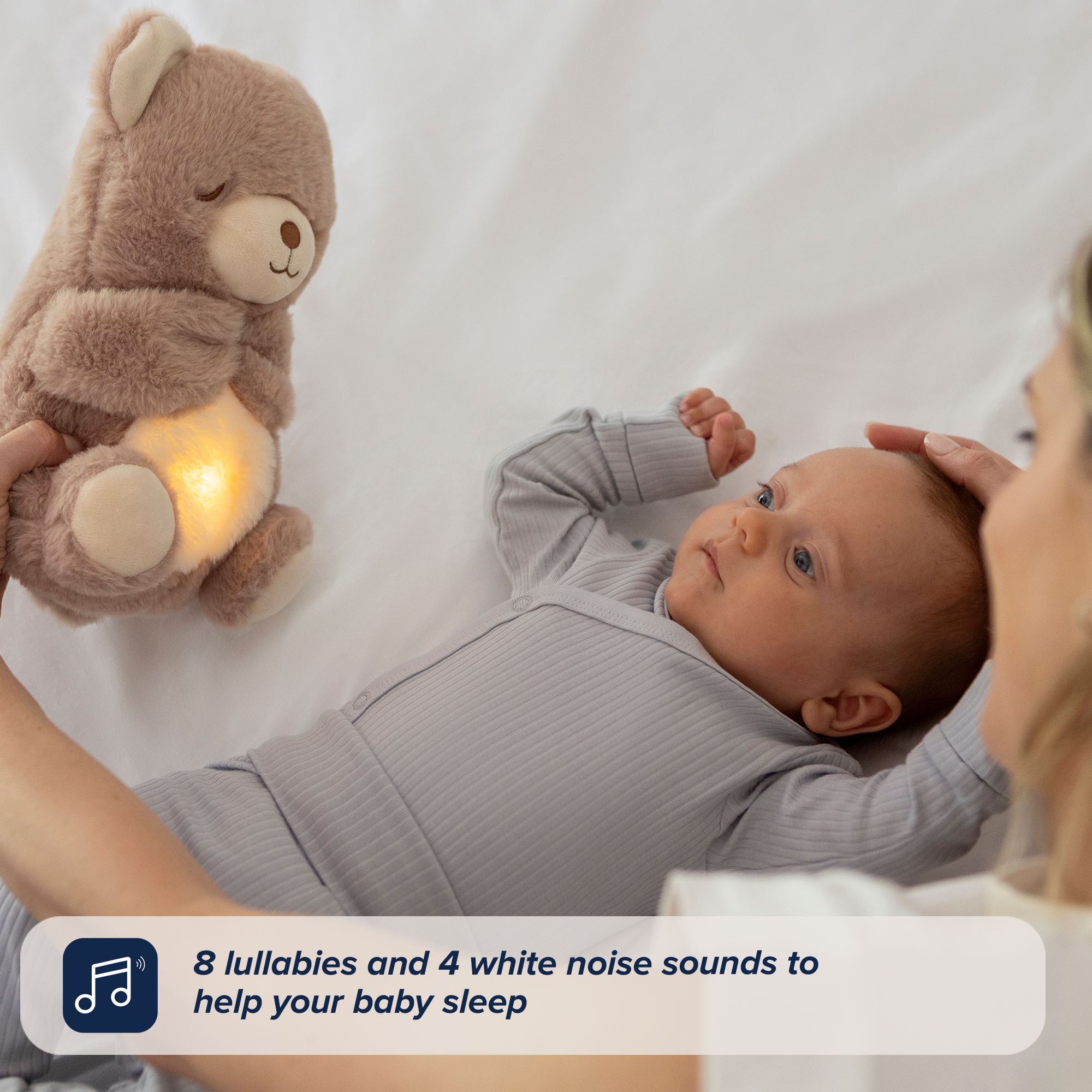 A baby in a gray onesie lies on a bed, gazing at an adult holding the Biscuit Bear Calming Comforter plush with glowing light. Text overlay reads: 8 lullabies and 4 white noise sounds make this comforter the perfect baby sleep aid.