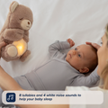A baby in a gray onesie lies on a bed, gazing at an adult holding the Biscuit Bear Calming Comforter plush with glowing light. Text overlay reads: 8 lullabies and 4 white noise sounds make this comforter the perfect baby sleep aid.