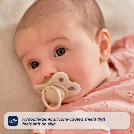 A baby in a pink outfit lies on a soft surface, using the RapidSoother Dummy & Pod with a beige silicone pacifier. Text overlay: “Hypoallergenic silicone-coated shield—gentle on baby’s skin.”.