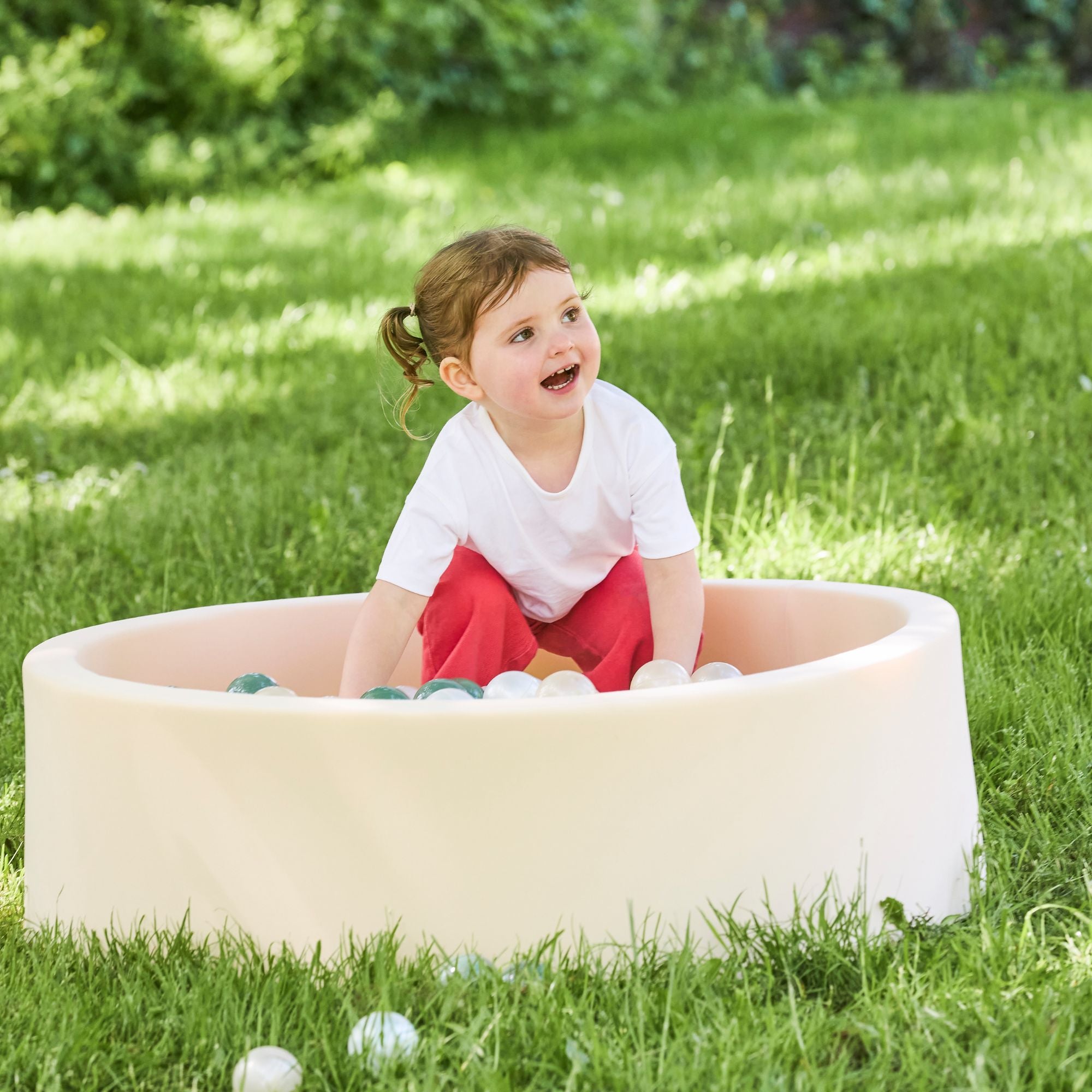 A young child with brown hair enjoys sensory play and develops hand-eye coordination while smiling in a Beige Ball Pit filled with plastic balls on a grassy lawn outdoors.