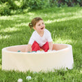 A young child with brown hair enjoys sensory play and develops hand-eye coordination while smiling in a Beige Ball Pit filled with plastic balls on a grassy lawn outdoors.