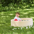 A young child with brown hair enjoys sensory play in a Beige Ball Pit outdoors on green grass, surrounded by scattered plastic balls and lush greenery.