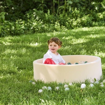 A young child with brown hair enjoys sensory play in a Beige Ball Pit outdoors on green grass, surrounded by scattered plastic balls and lush greenery.