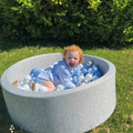 A young child with curly red hair, dressed in light blue, smiles and plays outside in a Nuby Baby Ball Pit filled with 200 blue & white balls, with green bushes in the background on a sunny day.