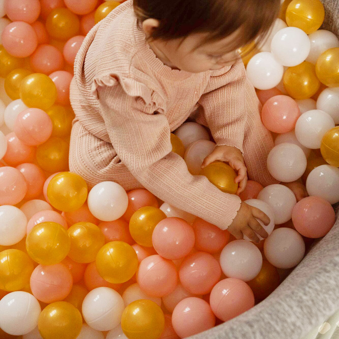 A ball pit for babies with pink gold and hite balls inside being played with by a baby girl.