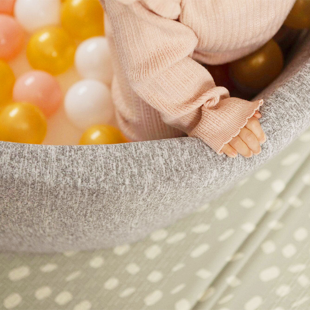A baby holding onto a ball pit for babies, with pink gold and white balls inside. This product is by Nuby.