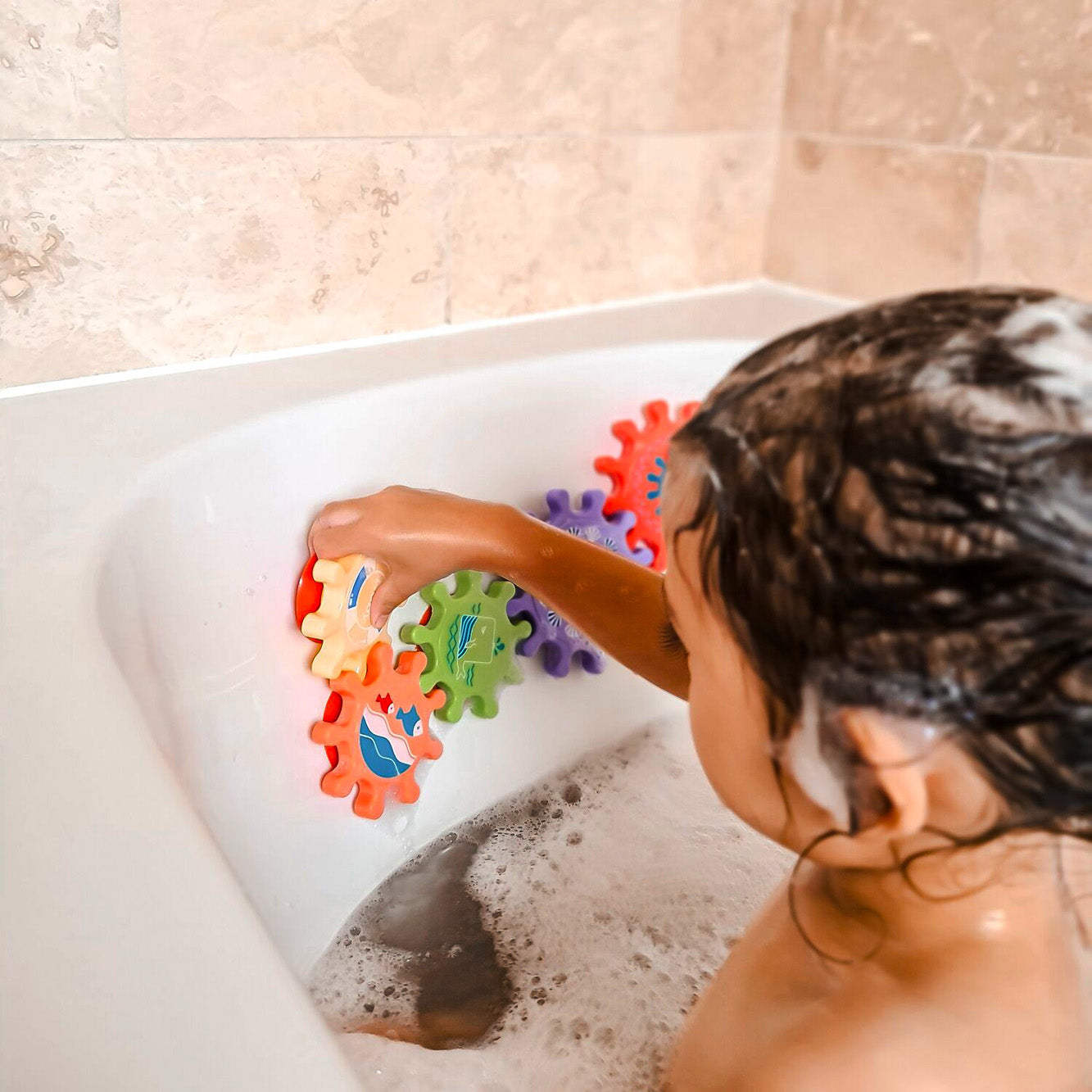 A child with wet hair and soap on their head plays in the bath, sticking Nuby Bath Toy Cog Set pieces to the tub wall for fun above-water play that helps develop motor skills.