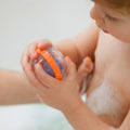 A young child sits in the bath, holding the Nuby Floating Bubbly Buds Bath Toy with an orange ring. Their hands and upper body are wet and covered in soap bubbles.