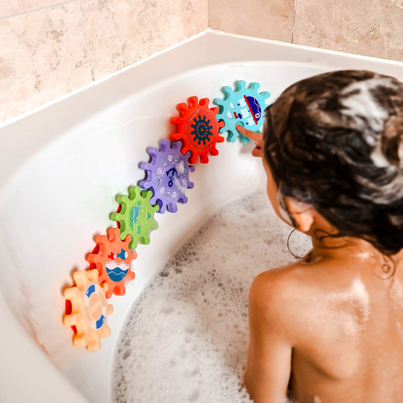 A child enjoys a bubble bath while playing with the Nuby Bath Toy Cog Set, spinning colorful gears attached to the side of a white tub and building motor skills.