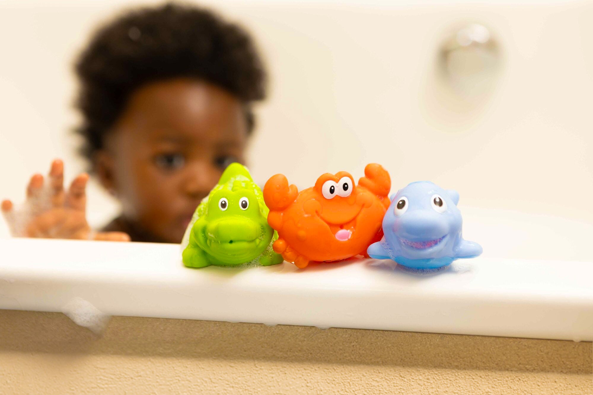 A toddler reaching for bath toys sat on the edge of a bath. These include a crocodile, crab and fish style character in vibrant colours.