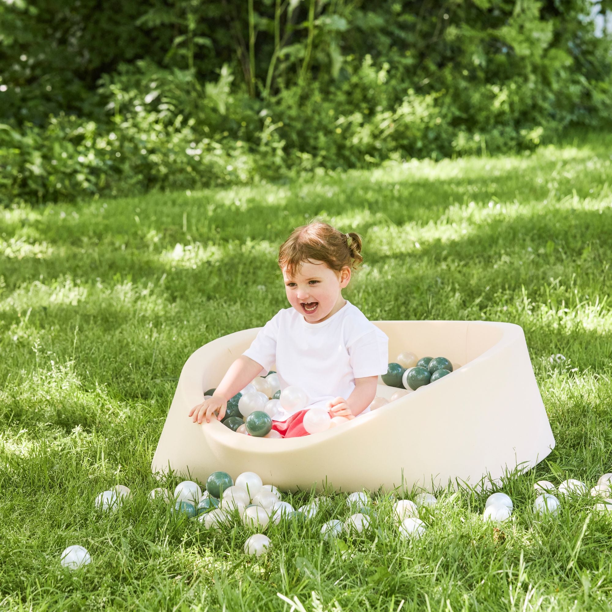 A young child with brown hair smiles while playing in a Beige Ball Pit on green grass outdoors, enjoying sensory fun with plastic balls. Lush greenery can be seen in the background.