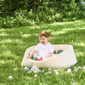 A young child with brown hair smiles while playing in a Beige Ball Pit on green grass outdoors, enjoying sensory fun with plastic balls. Lush greenery can be seen in the background.