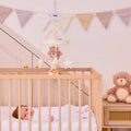 A baby lies in a wooden crib with white bedding. The Biscuit Bear Musical Cot Mobile hangs above. A brown teddy bear and rolled towel sit on a dresser nearby, with pastel bunting decorating the wall in the background.