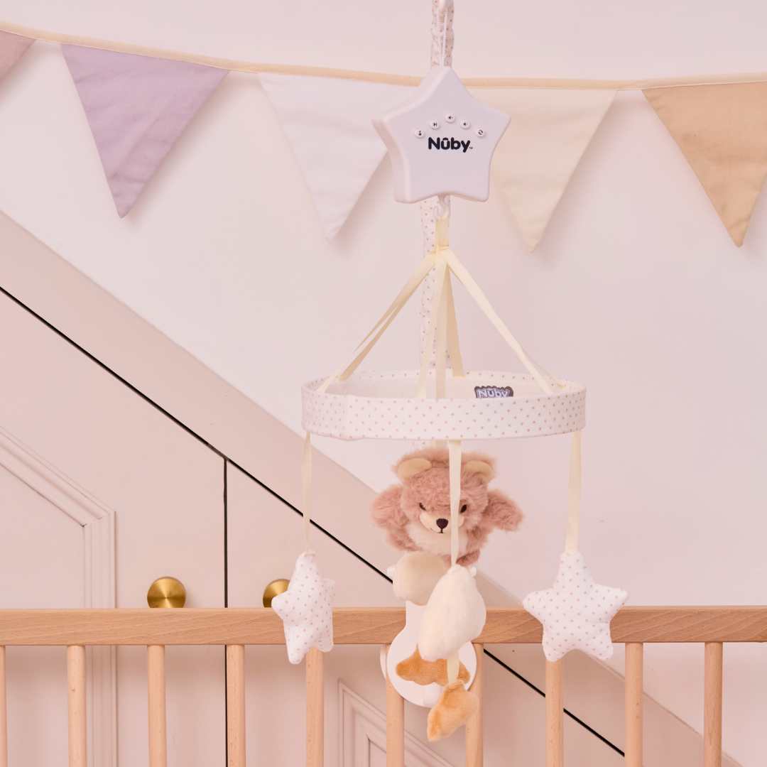 The Biscuit Bear Musical Cot Mobile, featuring plush stars and a teddy bear, hangs above a wooden crib. Pastel triangular bunting in the background adds charm to this cozy nursery scene.