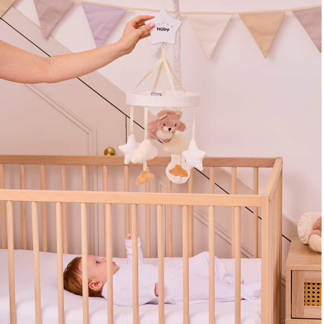 A baby lies in a wooden crib, gazing up at the Biscuit Bear Musical Cot Mobile with stars as an adult hand holds it. Soft pastel bunting decorates the wall in the background.
