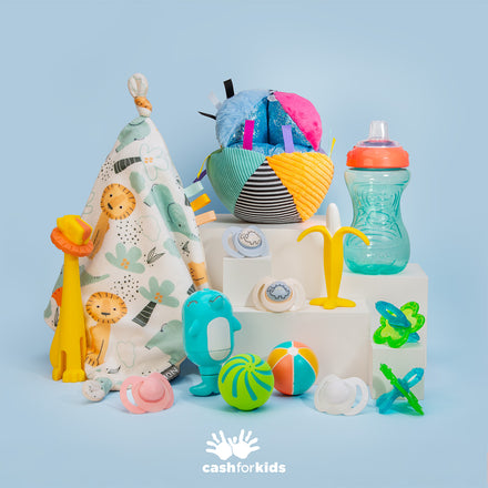 A vibrant arrangement of must-have baby essentials from the Nuby Baby Mystery Box—including baby toys, pacifiers, a bottle, a giraffe plushie, a patterned cloth, and more—are displayed on white blocks featuring the cashforkids logo against a light blue background.