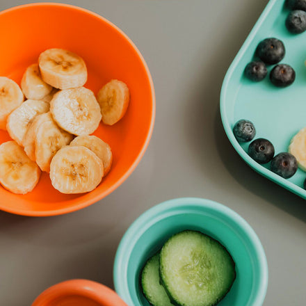 Sliced bananas in Nuby Baby Bowls 2 Pack, cucumber slices in a teal cup, and blueberries with banana slices on a teal plate are arranged on a gray surface.
