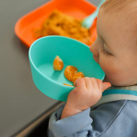 A baby holds a blue Nuby Baby Bowl (2 Pack), dishwasher safe, filled with orange fruit segments near their mouth, while an orange plate with food sits on the table in the background.