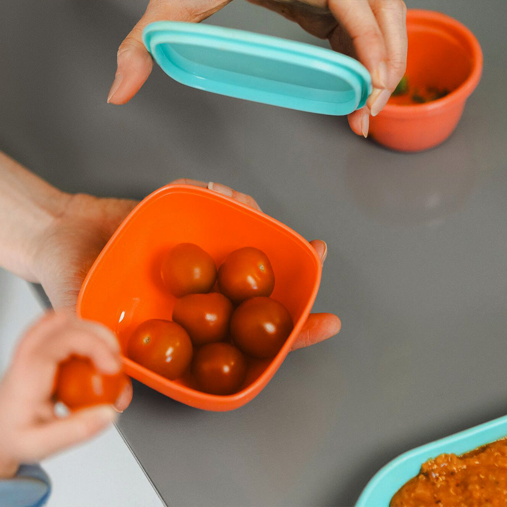 A pot of tomatoes by Nuby in an orange colour, this is part of their Brights Tableware range. Two hands are interacting with the pot, opening the lid for their child.