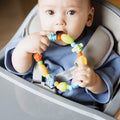 A baby in a high chair, dressed in blue, is holding and chewing on a colorful ring-shaped teether from the Nuby UK Teething Toy Set Bundle.