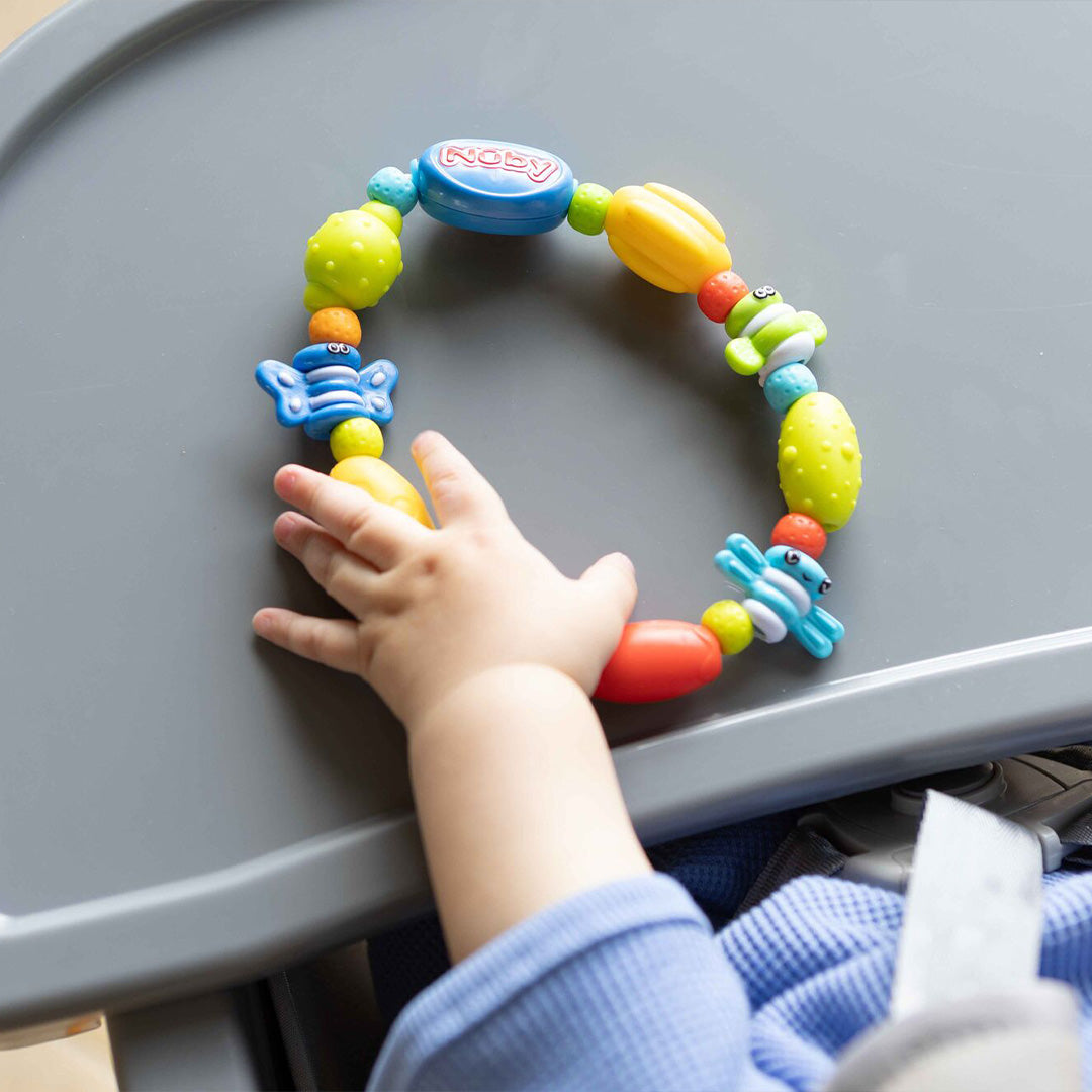A baby in a high chair reaches for a colorful teether from the Nuby UK Teething Toy Set Bundle, which features various shapes and textures.