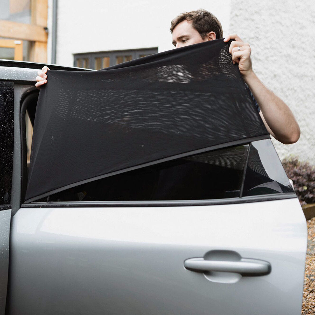 A person installs Nuby UK Car Window Shades (2 pack) on the rear window of a silver car parked by a white building, partially covering the open window to ensure UV protection for passengers.