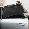 A person installs Nuby UK Car Window Shades (2 pack) on the rear window of a silver car parked by a white building, partially covering the open window to ensure UV protection for passengers.