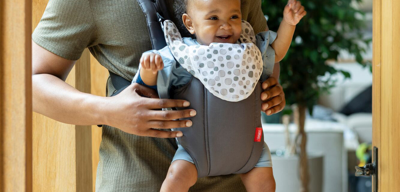 A baby being held in a 2 in 1 aby carrier by his Father while in front of an open light brown door.