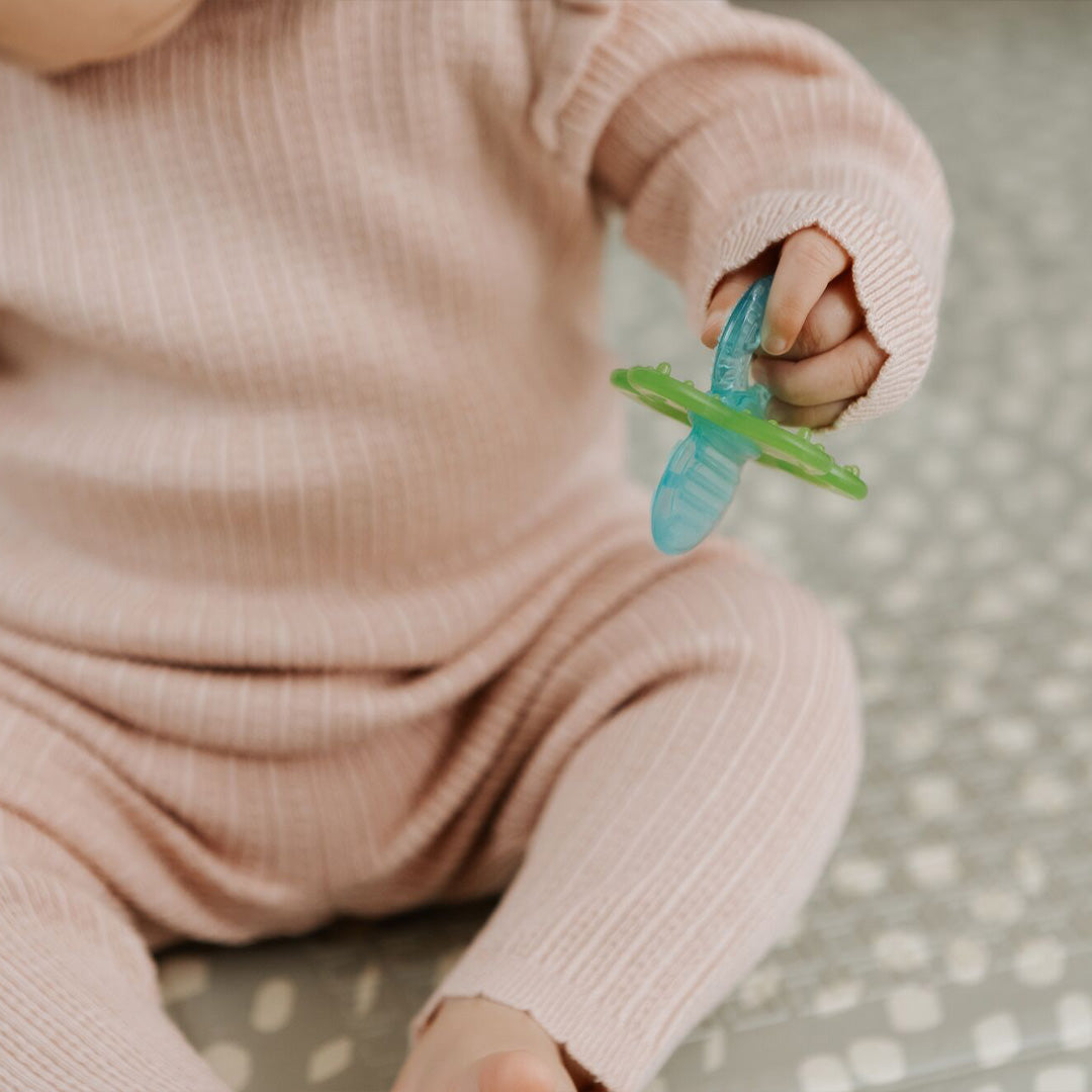 A baby in a pink ribbed outfit sits on a patterned surface, holding a blue and green pacifier from the Nuby UK Teething Toy Set Bundle.