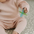A baby in a pink ribbed outfit sits on a patterned surface, holding a blue and green pacifier from the Nuby UK Teething Toy Set Bundle.