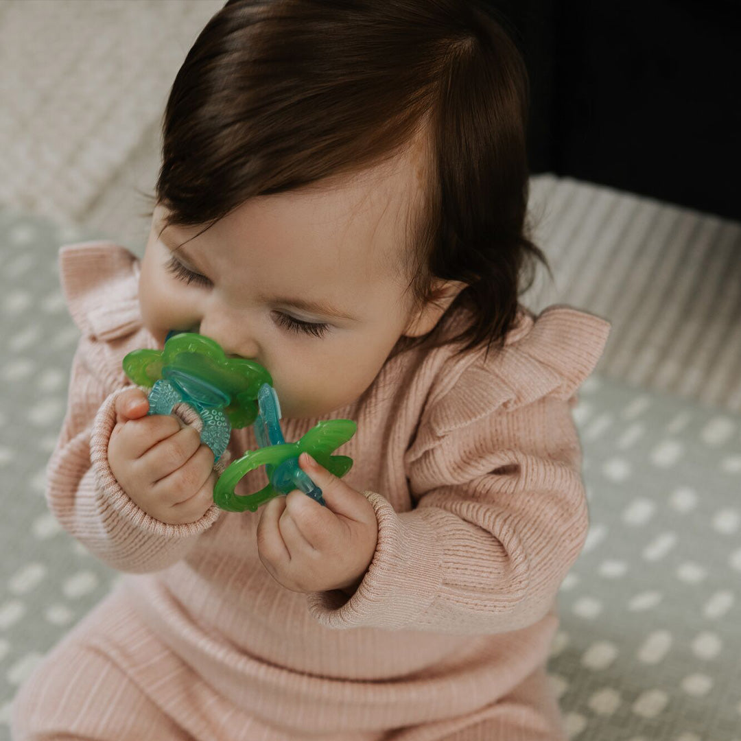 A brown-haired baby in a pink ribbed outfit sits on a patterned mat, holding the Nuby UK Teething Toy Set Bundle and bringing it to their mouth.