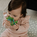 A brown-haired baby in a pink ribbed outfit sits on a patterned mat, holding the Nuby UK Teething Toy Set Bundle and bringing it to their mouth.