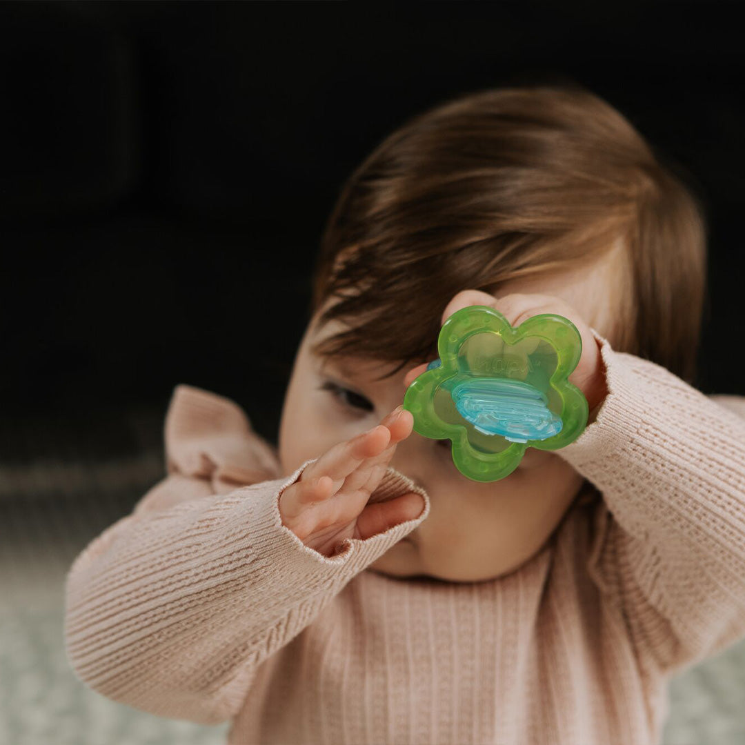 A baby in a light pink sweater holds the Nuby UK Teething Toy Set Bundle, a green flower-shaped toy, up to their eye while raising their other hand and partially covering their face.