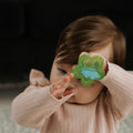 A baby in a light pink sweater holds the Nuby UK Teething Toy Set Bundle, a green flower-shaped toy, up to their eye while raising their other hand and partially covering their face.