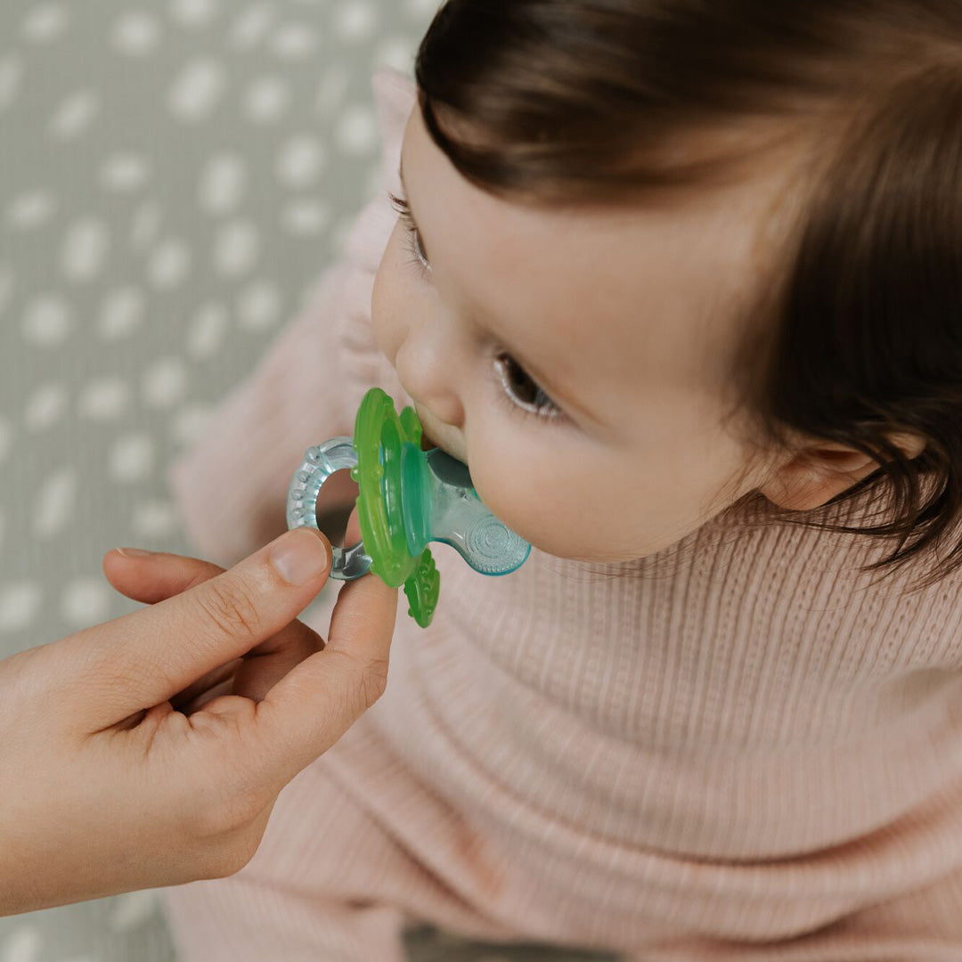 A baby in a pink sweater sits on a patterned surface while an adult offers a green and blue pacifier from the Nuby UK Teething Toy Set Bundle.