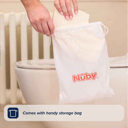 A hand places the Foldable Toilet Seat into a white Nuby drawstring bag by the toilet, highlighting its handy storage bag feature.