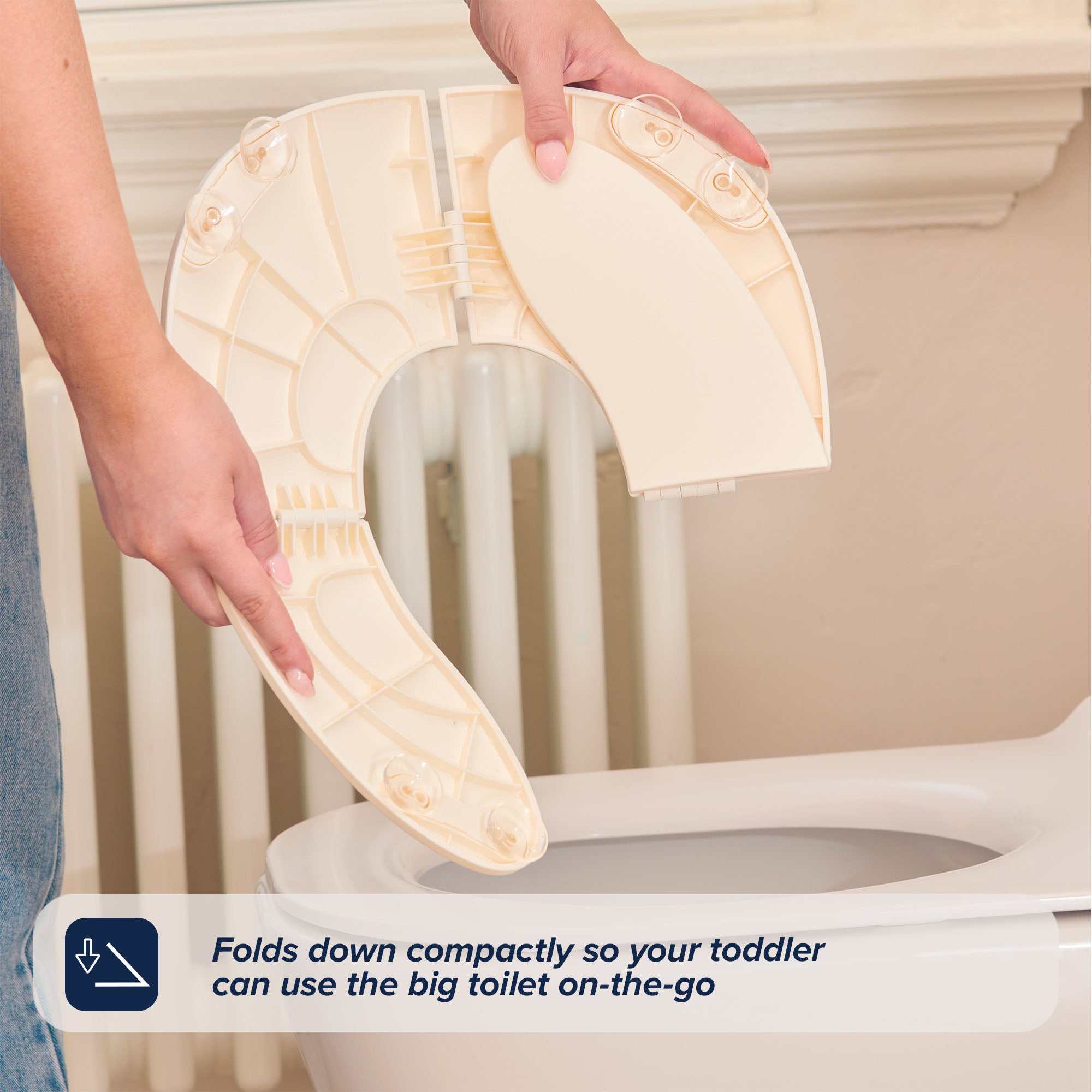 A person folds a light beige Foldable Toilet Seat above a toilet. The compact design lets your toddler use the big toilet anywhere. Text overlay: Folds down compactly for on-the-go toddler use.
