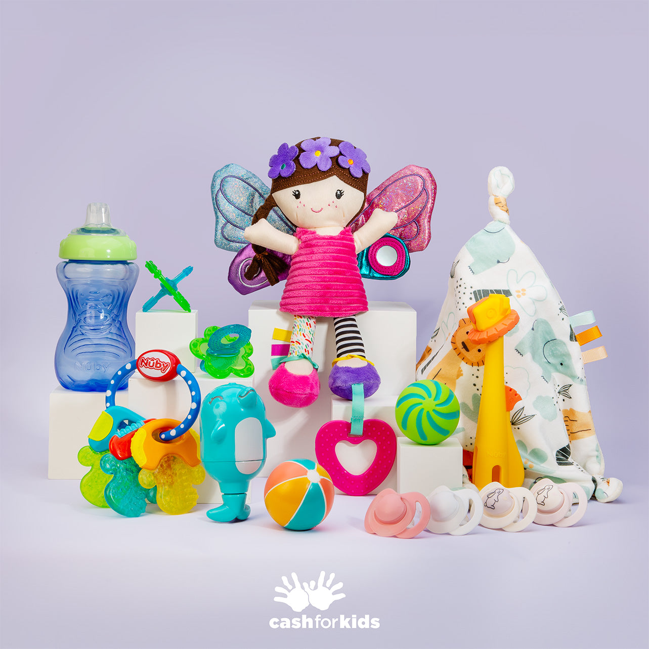 Discover the Nuby Mystery Box—a vibrant assortment of baby essentials featuring a winged doll, sippy cup, rattles, pacifiers, plush giraffe toy, baby ball, and a patterned blanket. Each item is thoughtfully arranged on a pale surface adorned with the cashforkids logo, making it the perfect choice for anyone seeking top parenting products.