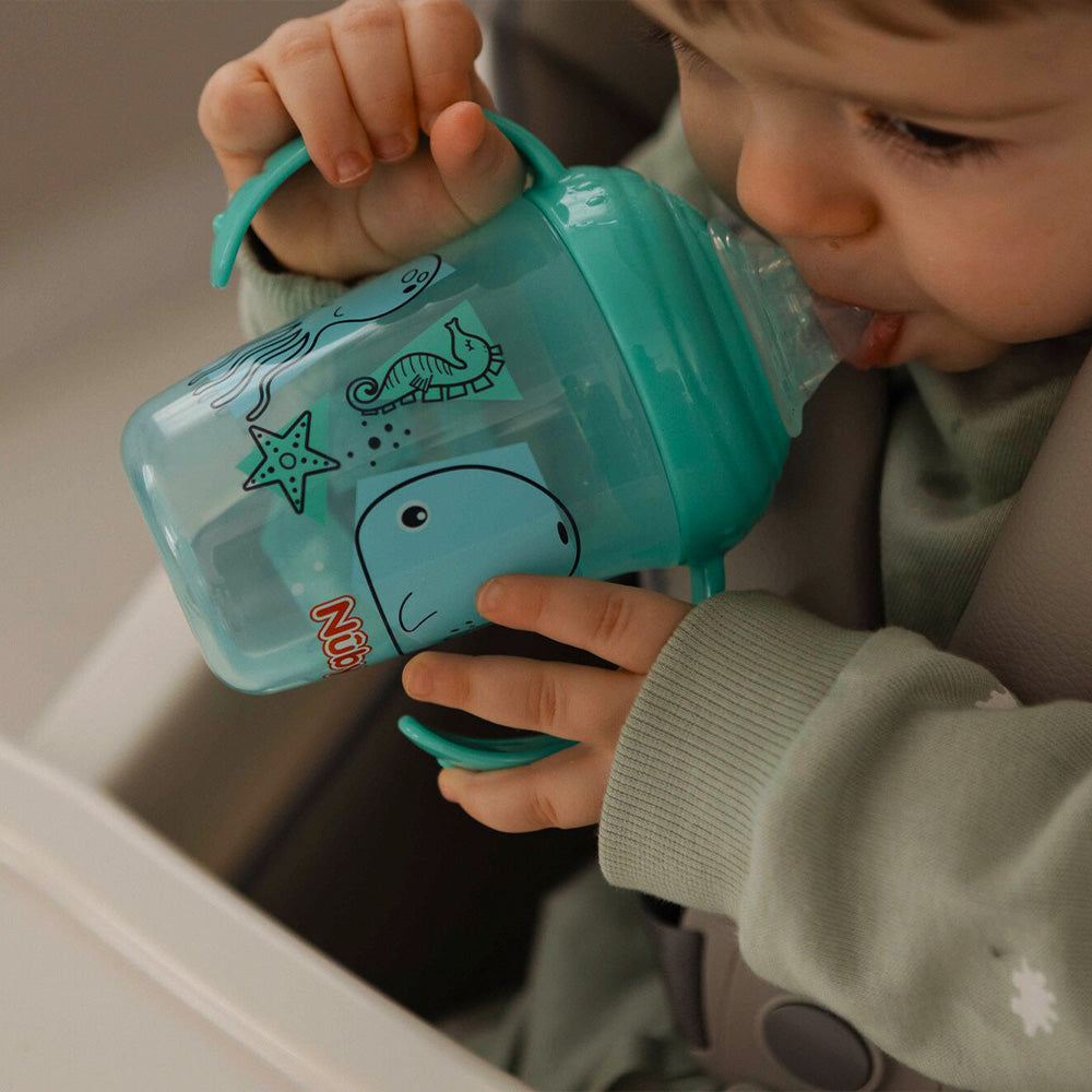 A young child holding onto the Grip n Sip cup by Nuby. It is green and blue with whales and sea animals on the sides of the cup.