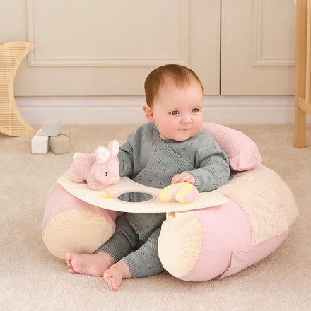 A baby in a grey knit outfit sits in the soft, pink and cream Little Hops & Raindrops Sit Up Seat, holding a plush toy and soft ball. Beige carpet, toy blocks, and a wooden shelf are in the background.