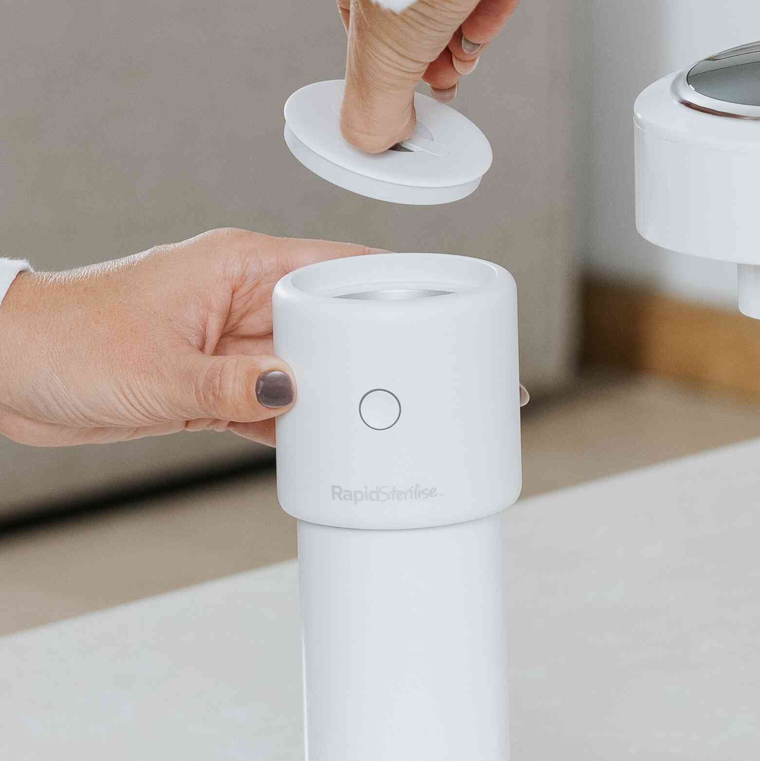 A person’s hand holds a white cylindrical device from Nuby UK’s Formula Making Bundle, while another lifts its round lid to reveal an opening at the top. The background is softly blurred.