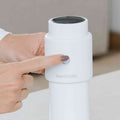 A hand with painted nails presses a button on the white cylindrical Nuby UK Formula Making Bundle device, which is part of a bottle feeding system, on a light-colored indoor surface.