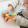 A baby in a textured gray sweater and bib sits in a high chair, holding the Nuby UK Teether Bundle featuring colorful rings and textured surfaces.