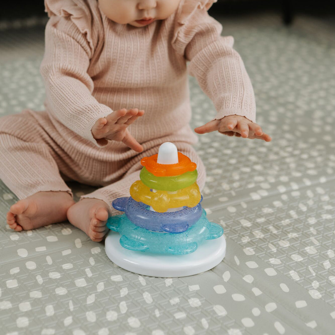 A baby in a pink outfit sits on a patterned mat, reaching for Nuby’s Icy Bite Stacking Rings—a colorful toy that also works as a teether.